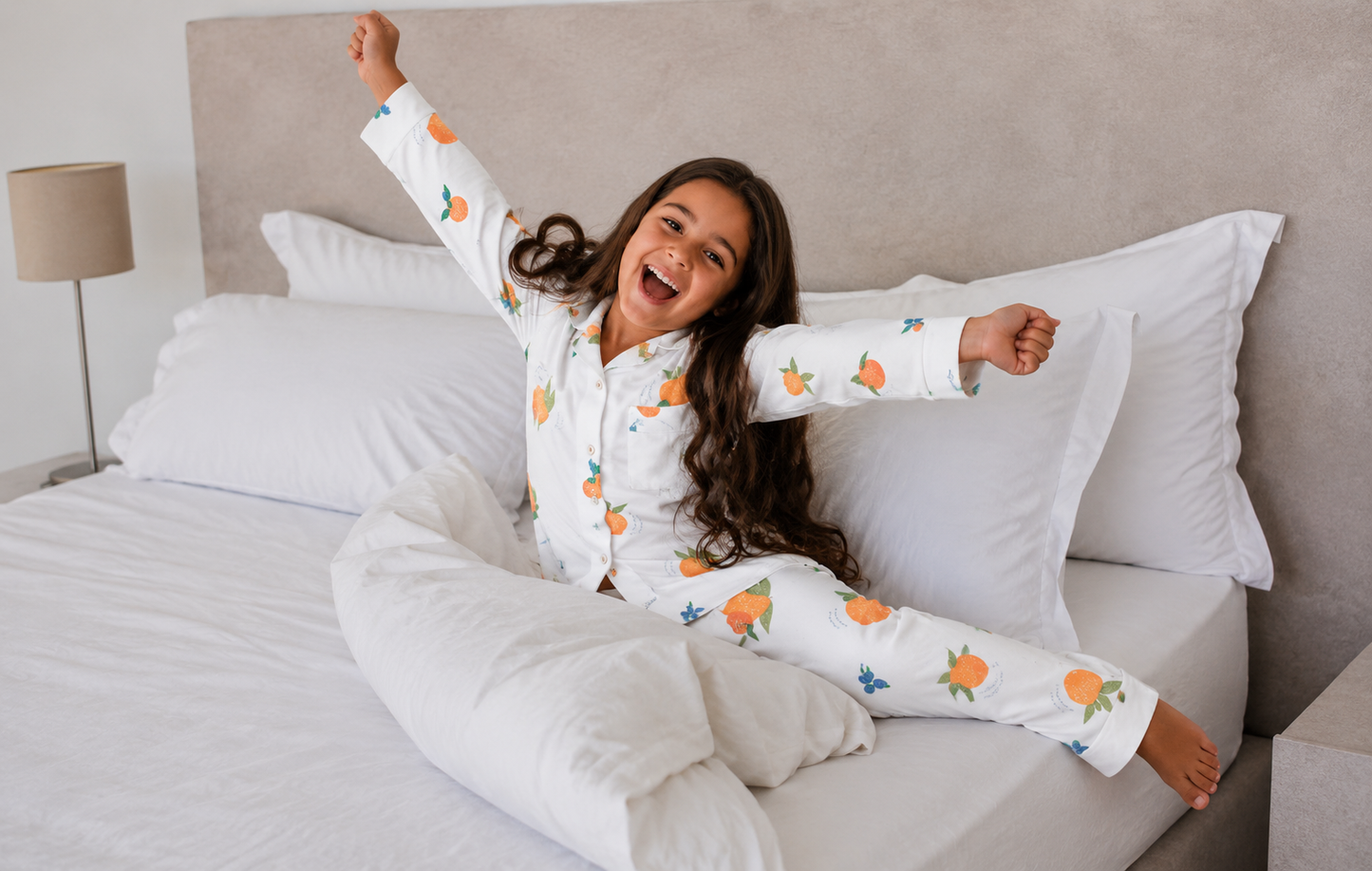 A young girl with long dark hair sits happily on a bed with white bedding, wearing a white long-sleeve pajama set printed with orange fruits and small blue butterflies.