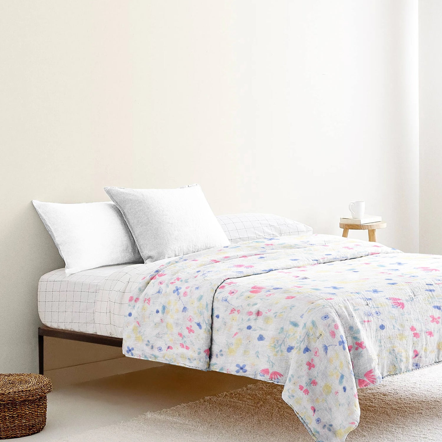 A bed with a white and multicolored floral print blanket, with two white pillows on it, placed in a room with light walls and a wooden floor.