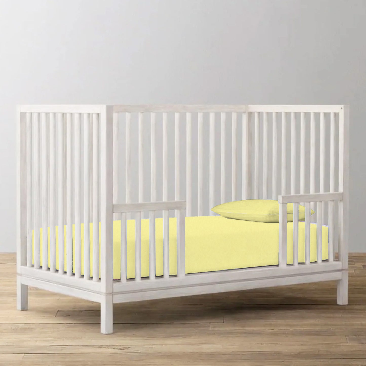 White crib with yellow bedding on a wooden floor against a gray wall