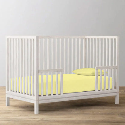 White crib with yellow bedding on a wooden floor against a gray wall
