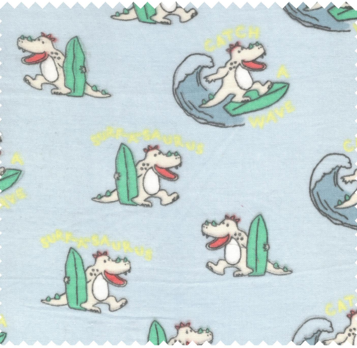 A close-up of a light blue fabric with a repeating cartoon dinosaur surfing pattern on a wave, with yellow text reading 'CATCH A WHALE'.