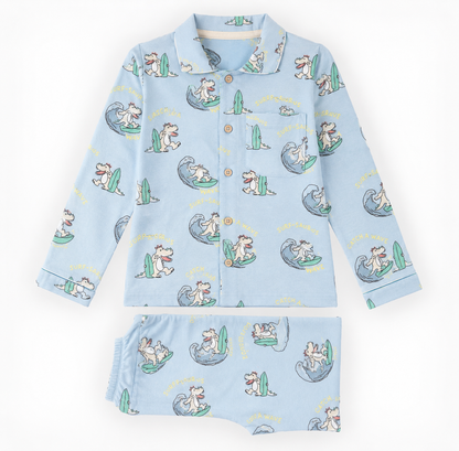 A two-piece children's pajama set laid flat on a white background, featuring a light blue long-sleeve button-front top and matching shorts with an all-over cartoon dog surfing pattern.