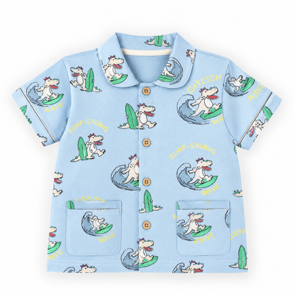 A light blue short-sleeve button-up shirt with an all-over cartoon dog surfing print, featuring a collar, gold buttons, and two front patch pockets on a white background.