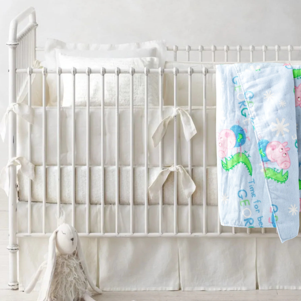 A receiving blanket with a Peppa Pig character pattern is draped over a crib with a pillow, a plush toy, and a framed picture of a sheep in the background.